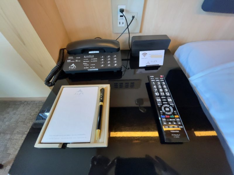 ホテルアルザ京都のフロントにつながる電話、テレビのリモコン、メモ帳、ボールペン、時計、Wi-Fiのパスワードが書かれた紙
